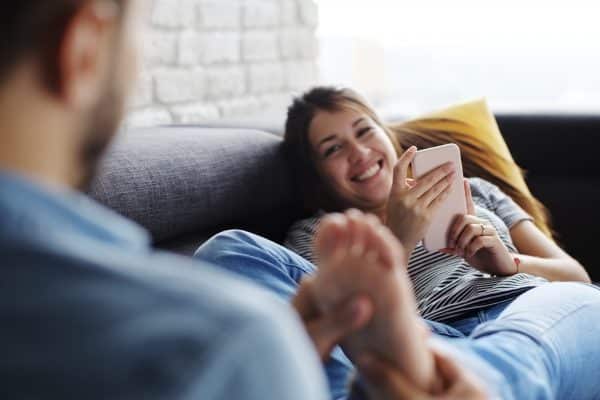 Young couple laying on couch at home.
The wife is using a mobile phone while the husband is giving her a foot massage which he learned from a couples massage course.